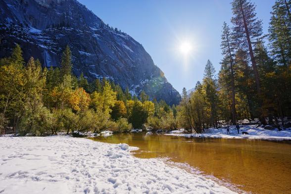 A sunlit riverbank covered in snow, surrounded by golden and green trees, with mountains in the background.