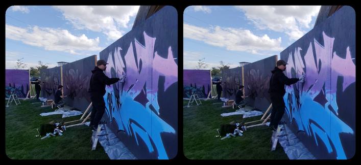 Black plywood walls sweep back from the right side of the image. In front of the closest wall, on a step ladder, is a graffiti artist, working in purples and blues. Blue sky beyond with clouds that compliment the letterforms.