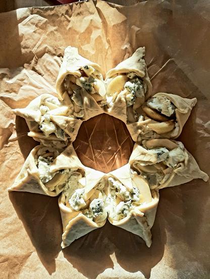 Ein sternförmiges Hefeteig-Gebäckstück mit einem Loch in der Mitte. In den Zacken ist eine Füllung aus Birnenschnitze und Gorgonzola-Bröckchen.
Das Ganze liegt auf Backpapier und zur Hälfte im Sonnenlicht.