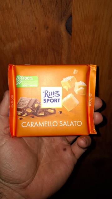 Parts of a hand are visible, holding an orange square Ritter Sport chocolate bar with “Caramello Salato” written on it. In the background, wooden boards of an arolla pine door.