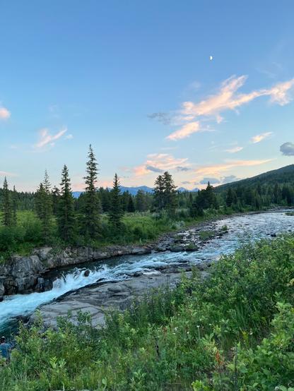 A scenic riverside view featuring a rushing river surrounded by rocky outcrops and lush greenery. Pine trees line the banks, with mountains in the background and a clear blue sky adorned with soft clouds and a crescent moon.