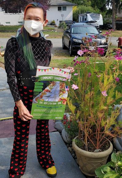 Me, with rainbow hair and a face mask wearing a black patterned top and heart-patterned jumpsuit, holding a poster for a community fruit-sharing event, with pink flowers in a pot and houses in the background.