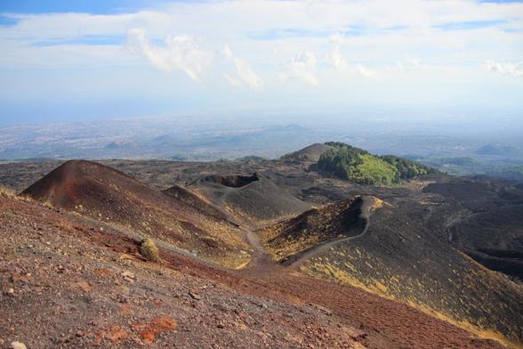 A panoramic view of a volcanic landscape. In the foreground, a slope of reddish, stony terrain, dotted with patches of dry vegetation. The center of the image shows several dark-colored volcanic cones, some with visible craters and winding paths. The surrounding terrain is covered in rugged, black volcanic rock. Towards the right, a hill stands out with its dense vegetation of green trees, offering a contrast to the arid landscape. On the distant horizon, a vast plain housing a coastal town can be seen, blurred under a blue sky with scattered clouds