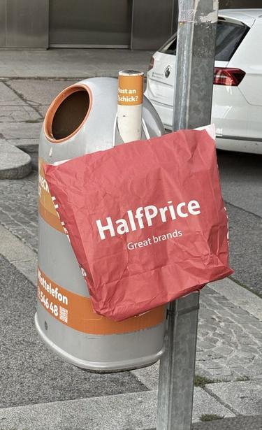 Mistkübel auf der Strasse. An diesem hängt eine rote Einkaufstasche mit dem Aufdruck "HalfPrice Great brands"