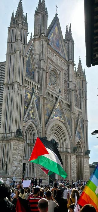 L'immagine mostra una folla di persone radunate davanti a una maestosa cattedrale gotica, caratterizzata da archi acuti, torri spiccate e mosaici colorati. Al centro della scena, una bandiera palestinese è visibile, sventolando con orgoglio. In primo piano, si notano anche una bandiera arcobaleno, simbolo della comunità LGBTQ+, e un cartello bianco, probabilmente con un messaggio di protesta. La cattedrale, con la sua architettura imponente e dettagli intricati, è un simbolo di storia e cultura. La presenza di diverse bandiere suggerisce un evento di protesta o manifestazione, con la folla che riempie le scale e le aree circostanti.