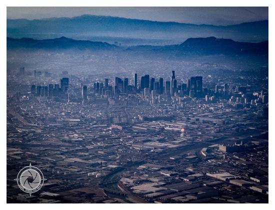 A shallow smog blankets the Los Angeles basin with the skyscrapers of downtown LA rising above in the foreground of the picture. Beyond the skyscrapers, haze obscures the details of the urban sprawl. The mountains to the north of the city appear silhouetted in the background.