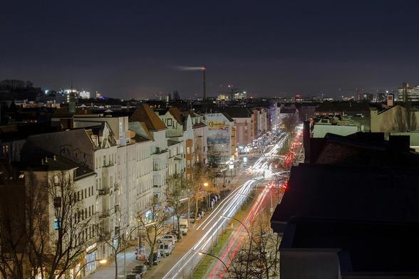 Nachtaufnahme einer Hauptstraße in Berlin-Wedding mit beleuchteten Wohnhäusern, Reklametafeln und Lichtspuren des Verkehrs.