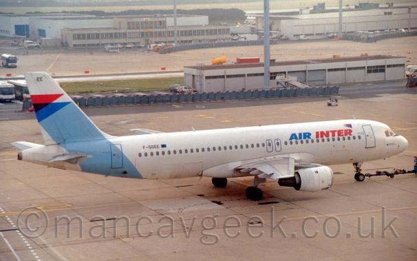 Elevated side view of a twin engined jet airliner being towed from left to right by an out-of-frame tow truck attached to the nose-wheel buy a long black pole.
The plane is largely white, with blue and red "Air Inter" titles on the upper forward fuselage, and the black registration "F-GGEE" next to a small EU flag on the upper rear.
There is a wide, light blue band around the rear fuselage and tail, with red and dark blue triangles at the top of the tail, along with the letters "EE" right at the top.
Grey concrete apron fills the foreground and much of the background, with low, white buildings in the background, and tall lighting towers reaching out of the top of the frame.