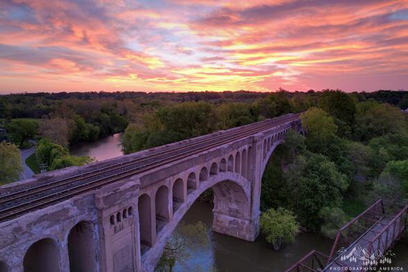 A view from a drone of a sunrise with a cement railroad bridge spanning a river in the foreground.