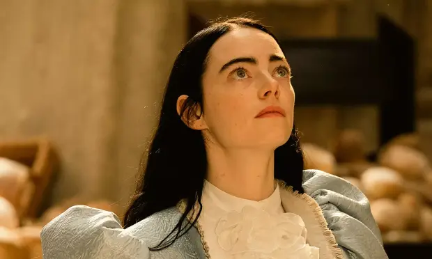 Fotograma de la película donde se ve en primer plano a Emma Stone, en el papel de la protagonista, mirando hacia arriba, seria, en actitud contemplativa. Va vestida con un traje "de época" azul celeste y blanco. Va peinada con una melena morena y lacia. La luz es amarillenta, el fondo está difuminado.