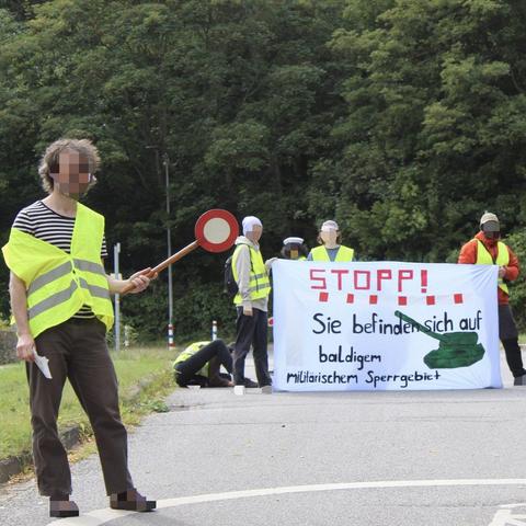 Im Hintergrund halten vier Menschen ein Transpi auf dem "Stopp! Sie befinden sich auf baldigem militärischen Sperrgebiet" drauf steht. Auf dem Transpi ist ein Panzer gemalt. Im Vordergrund steht ein Mensch der eine rote Kelle zum Anhalten von Personen in der Hand hält. Alle Menschen tragen gelbe Warnwesten.