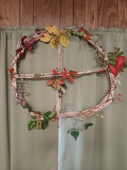 A wheel with a cross in the center made of reeds wrapped in grapevine. It's decorated with fall flowers, foliage, and fruit. Notable mentions: purple aster, goldenrod, the bright red leaves of sumac and Virginia creeper, a bundle of wild grapes in the center, and a pear and a honey mushroom hanging off the bottom.
