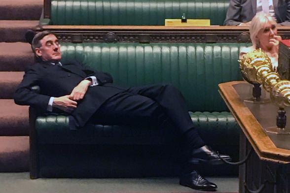 The image shows a man in a black suit and tie, reclining on a green leather bench in what appears to be a parliamentary chamber. He is lying on his back with his legs crossed and his head resting on the armrest, appearing to be asleep. The bench is part of a larger seating arrangement with green leather upholstery, and there is a wooden table with a golden object on it in the foreground. In the background, another person is seated, partially visible, wearing a light-colored outfit. The setting suggests a formal and official environment, likely a legislative assembly.
(It is the House of Commons)