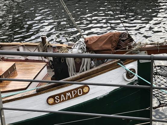 Durch ein Geländer ist das Holz-Namensschild auf weiß gestrichener Fläche am Holzboot. Wasser im Hintergrund dunkel mit Wind-Wellen.