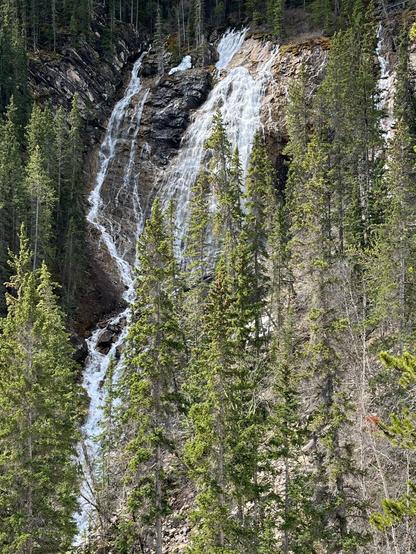 A waterfall cascading down rocky cliffs surrounded by lush evergreen trees. The scene captures a natural landscape with rushing water and a mix of greenery and rock formations.