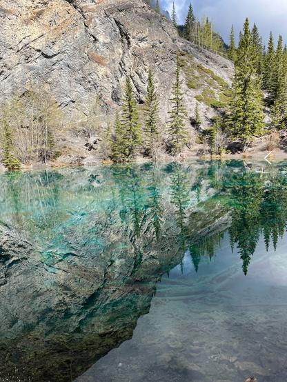 A serene scene featuring a clear, reflective body of water surrounded by rocky terrain and tall, green coniferous trees. The vibrant colors of the water reflect the forest and rocky landscape, creating a tranquil atmosphere.