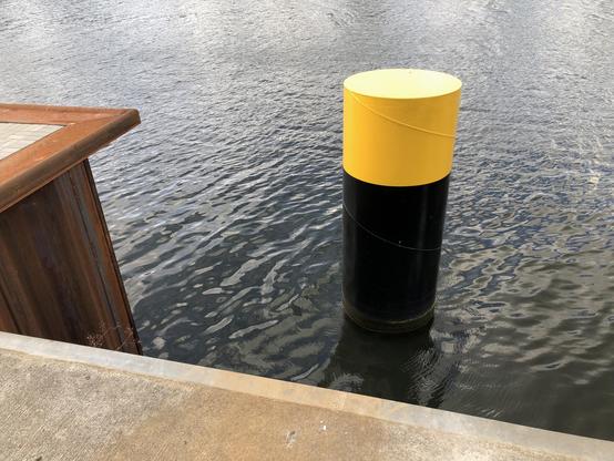 Eine Dalbe mit frischer Farbe, unten schwarz, oben gelb im krausen Wasser. Unten am Bildrand ein Stück Kaikante.