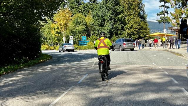 A cyclist on a two-lane paved road. There are cars and pedestrians in the background.