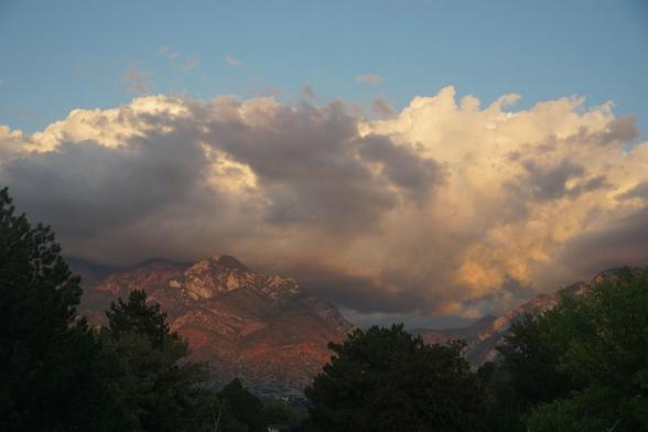 The Wasatch Front seen from my office window, looking East. In the foreground are trees, the mountains are also covered in trees. Their leaves are changing colours. The last rays of the sun tinge the ridges of the mountains. A thick layer of clouds, lit by the sunset, cloak the mountains. Above it all the sky is blue.