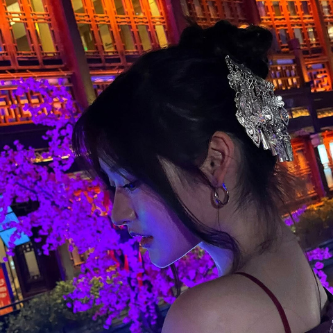@jemmesedi The image features a woman with her hair styled in an updo, adorned with a silver hair accessory that has intricate designs and dangling elements. She is wearing a hoop earring in her visible ear. The background showcases a traditional building with red wooden structures and illuminated windows, creating a warm ambiance. In the foreground, there are purple-lit trees, adding a vibrant contrast to the scene. The overall setting appears to be an evening event or a culturally themed location, with the lighting enhancing the aesthetic appeal.

Provided by @altbot, generated privately and locally using Ovis2-8B