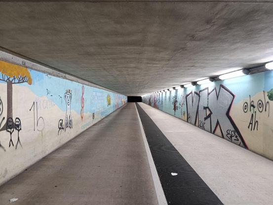 Eine Unterführung, beleuchtet am Ende dunkel, an beiden Seiten Graffitis, die von Graffitis übermalt worden sind.
Unten ein Kanal mit Metallgitter überdeckt..