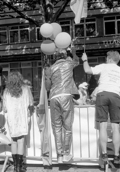 Three people seen from behind near a metal barrier; one in a shiny patterned suit holding balloons, another in a white T-shirt and shorts holding a flag, and a third in a light dress and boots.