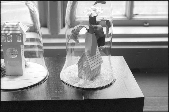 A black and white photograph of small houses inside glass domes on a bench, in a window. Part of an exhibition of paper art.