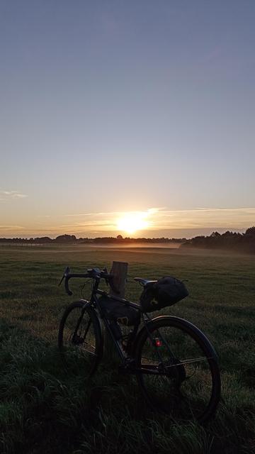 Ein Gravelbike mit Rahmen- und Satteltaschen lehnt an einem Weidezaun. Im Hintergrund der Sonnenaufgang. Auf dem Feld steht etwas Morgendunst.