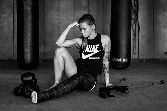 Kickboxerin auf den Matten der Boxhalle sitzend mit einem Bein komplett in schwarzem Gips. Neben sich Boxhandschuhe und Krücken.