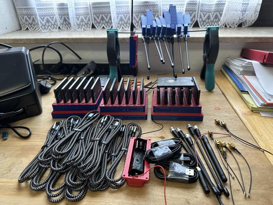 Auf einem Holztisch stehen mehrere rot-blaue Mehrfachladegeräte für T1000-E Meshtastic-Knoten, bestückt mit zahlreichen Funkmodulen. Links liegen ordentlich aufgewickelte, geflochtene USB-Kabel. Rechts sind Antennen, Adapter und Kabel zu sehen. Im Hintergrund befinden sich halbfertige Gehäuse für Heltec Tracker, bereits mit montierten Antennen. Außerdem stehen einige Werkzeuge wie große Klemmen auf dem Tisch.