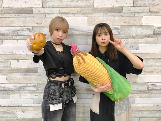 Two young women pose against a wooden plank wall background. The woman on the left holds up a potato while wearing a cropped lace top and high-waisted jeans. The woman on the right holds a giant corn prop and makes a peace sign with her other hand.
