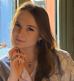 A young woman looking at the camera. She is white, with mouse-blonde hair, and her head is tilted towards the viewer with her right hand touching her chin: there is a bright window behind her.