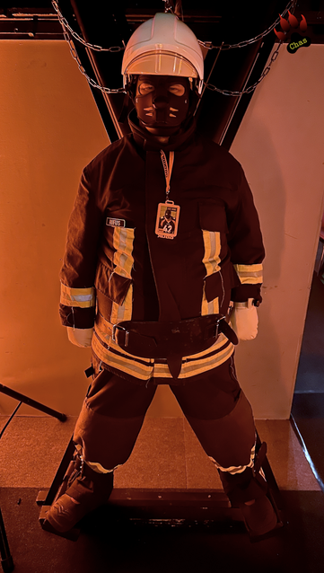 human pup, wearing a complete firefighter uniform, Haix boots, helmet and padded mitts, bound to a cross in a dimly lit playroom