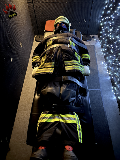 human pup, wearing a complete firefighter uniform, Haix boots, helmet and padded mitts bound to a bondage bench in a dimly lit playroom
