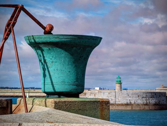 A close-up of a large, weathered, green-patinated bell sitting on a stone wall in the foreground. In the background, across the blue water of the harbour, a white stone pier leads to a small, green-domed lighthouse. The sky above is filled with fluffy white clouds against a blue sky.