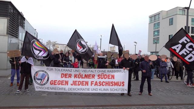 Das Bild zeigt eine Demonstration in Köln. Eine Gruppe von Menschen hält ein großes Banner mit der Aufschrift „Der Kölner Süden gegen jeden Faschismus. Für Toleranz und Vielfalt ohne Rassismus. Menschenrechte, Demokratie und Klima schützen.“ Eine AntiFa-Fahne mit dem Farbverlauf der Warming Stripes ist auf der linken Seite. Die Aufschrift lautet: Nazis leugnen die Gefahr - Klimaschutz ist ANtiFa. Mehrere Teilnehmer tragen schwarze AntiFa-Fahnen mit verschiedenen Texten wie Antifaschistische Aktion und FCKAFD.
