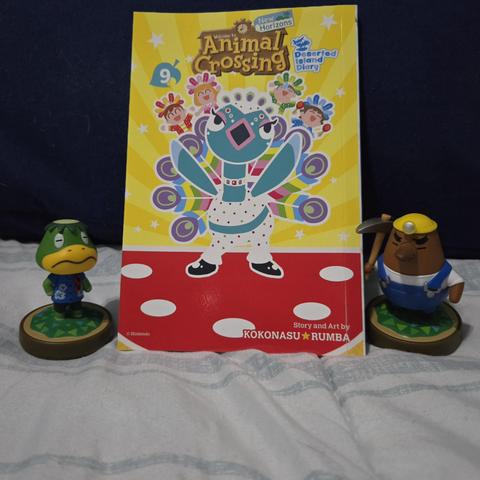 Photograph of Animal Crossing: New Horizons Deserted Island Diary Volume 9 sitting against a blue colored pillow. The cover is mostly yellow with the bright blue colored Pave (very colorful peacock) dancing in the center with tiny images of the main human cast behind them dressed in Festivale oufits. On either side of the book are Amiibo figures of Resetti (right) and Kapp'n (left).