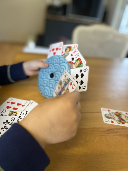 Runder Kartenhalter mit blauem Bogenmuster, in dem verschiede Spielkarten stecken. Eine Hand hält den Kartenhalter, die zweite Hand steckt eine weitere Karte in den Halter.