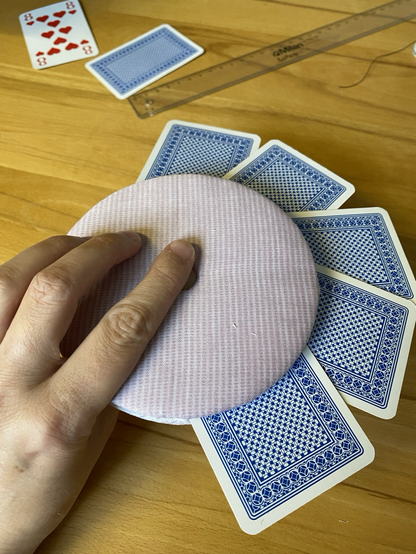 Runder Kartenhalter mit einem rosa Karomuster. In den Kartenhalten sind 5 Spielkarten gesteckt. Zu sehen sind nur die blau-weißen Rückseiten der Karten. Der Hintergrund ist ein Holztisch. Der Kartenhalter wird mit einer Hand festgehalten.
