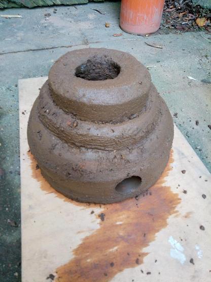 Auf einen Brett steht ein 3- etagiger Lehmofen, obem ein größeres Loch, unten für die Zuluft ein kleines. Sieht aus wie eine Hochzeitstorte.