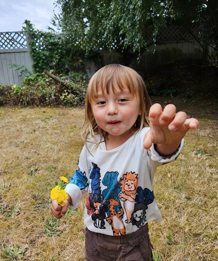A young child with fair hair, wearing a shirt decorated with animal cartoon characters and brown pants, stands in a backyard holding a bouquet of yellow dandelions in their left hand, reaching towards the camera with their right hand. The background shows a fence, vegetation, and a cloudy sky.
