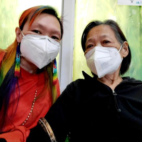 Two women wearing KN95 face masks, one with rainbow-colored hair and beaded earrings, the other with dark hair, looking towards the camera.