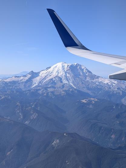 A picture taken from an airplane window cruising at peak level with Mt Rainier. the green covered hills, the fog, and the. the snowy peak of the volcano is breathtaking