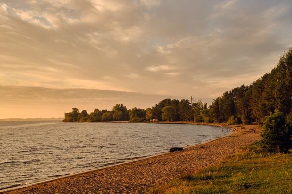A serene lakeside view during sunset, featuring a sandy beach, calm waters, and trees lining the shore. Gentle waves create a tranquil atmosphere beneath a sky filled with soft clouds.