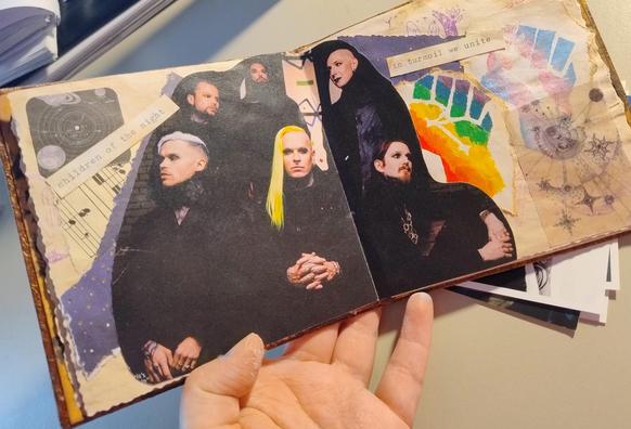 A photo of a small scrapbook in a hand. The main feature is a dark image of the band lord of the lost, with painted fists of solidarity in the original pride and trans pride colours, and stars. The lyrics say "children of the night / in turmoil we unite"