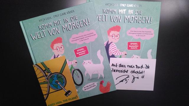 Auf dem Bild sind zwei Exemplare von Katja Diehls Buch "Komm mit in die Welt von morgen!" zu sehen. Zudem sind die, jedem Exemplar beigefügte Postkarte von Guido Kühn und die von Katja Diehl persönlich geschriebene Grußbotschaft abgebildet.