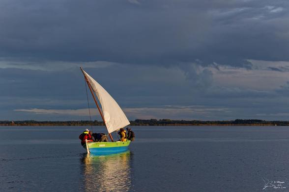 Photo d'une pinasse à voile bleu et verte, voile hissée qui s'éloigne et est éclairée par le soleil couchant, sur une mer d'huile bleu ardoise qui reflète un ciel couvert.
La photo est de Yo Photographie, Regard & Lumière