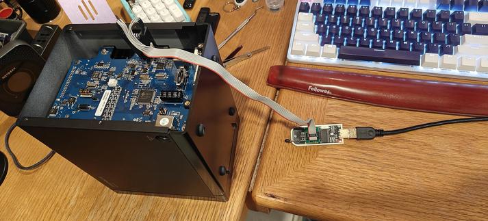 This is a photo of a technical project taking place on a wooden desk.

An open black electronic device, a Mediasonic Probox, has its internal blue circuit board exposed. A gray ribbon cable is connected from the circuit board to a small green USB programmer tool. This tool is then connected via a black USB cable to a computer.

In the background, a mechanical keyboard with blue and white keycaps, a wrist rest, and other desk items are visible. The setup suggests someone is in the process of programming, debugging, or modifying the firmware of the electronic device.
