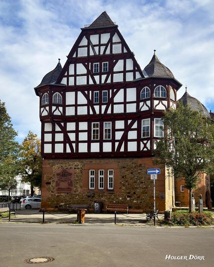 Ansicht des Fachwerkflügels des Neuen Schlosses in Gießen. Zwei runde Türme flankieren die reich verzierte Fachwerkfassade aus dem 16. Jahrhundert. Durch die leicht schräge Perspektive entsteht ein Eindruck von Schlosscharakter und historischer Pracht, verstärkt von einem wolkigen Himmel im Hintergrund.