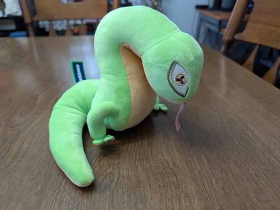 A plushie of Snecko, a green snake-like reptile with two small legs midway along its length, its body rearing up forward from the legs, before curving back down to have the head face down at an angle. Its belly is yellow, as are the irises of its eyes, with black pupils that split the iris into three segments. A pink forked tongue hangs out from its mouth.